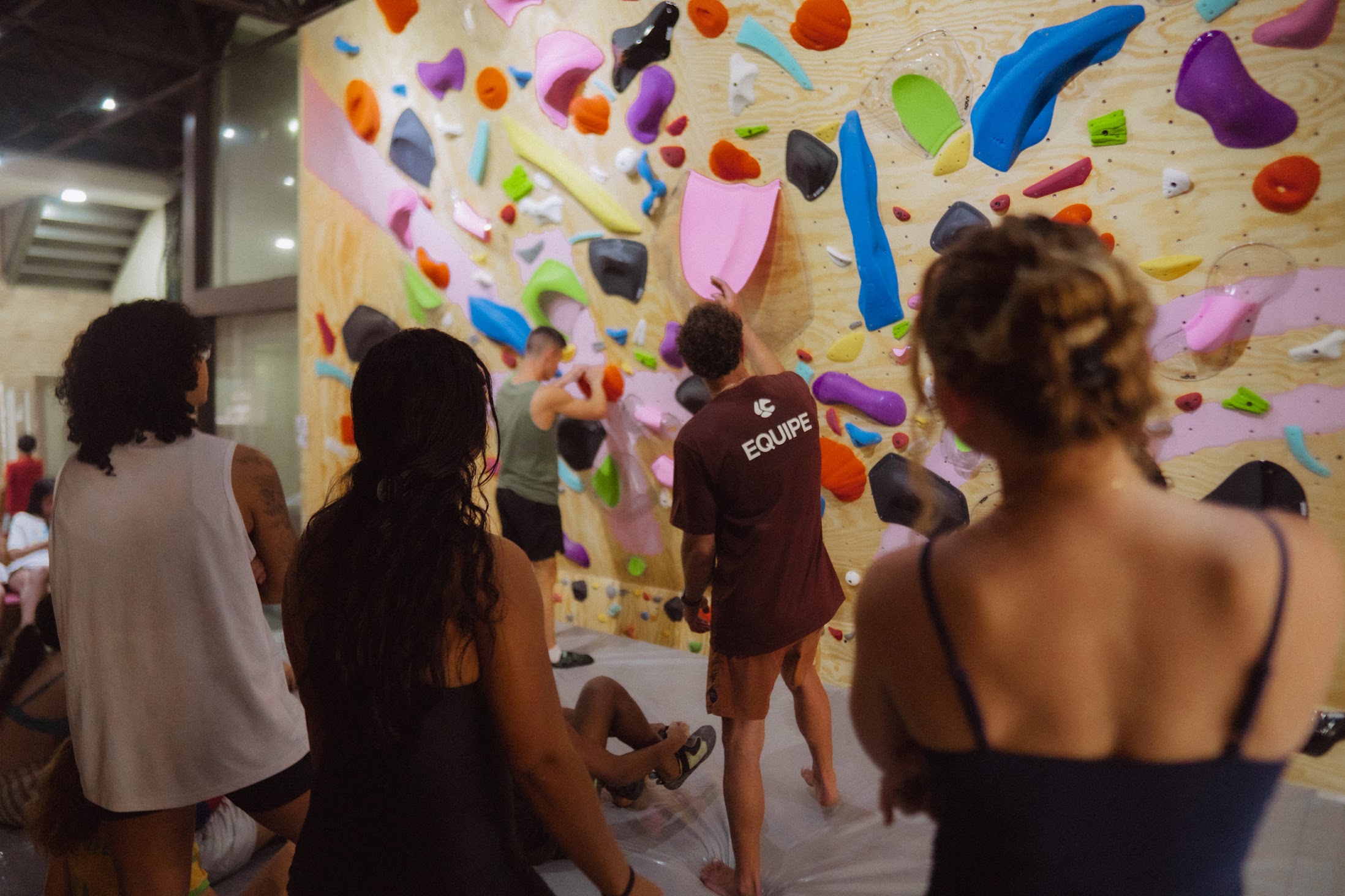 Grupo de pessoas de costas, em pé diante da parede de boulder, acompanhando quem escala; uma delas veste a camiseta da equipe da Colmeia.