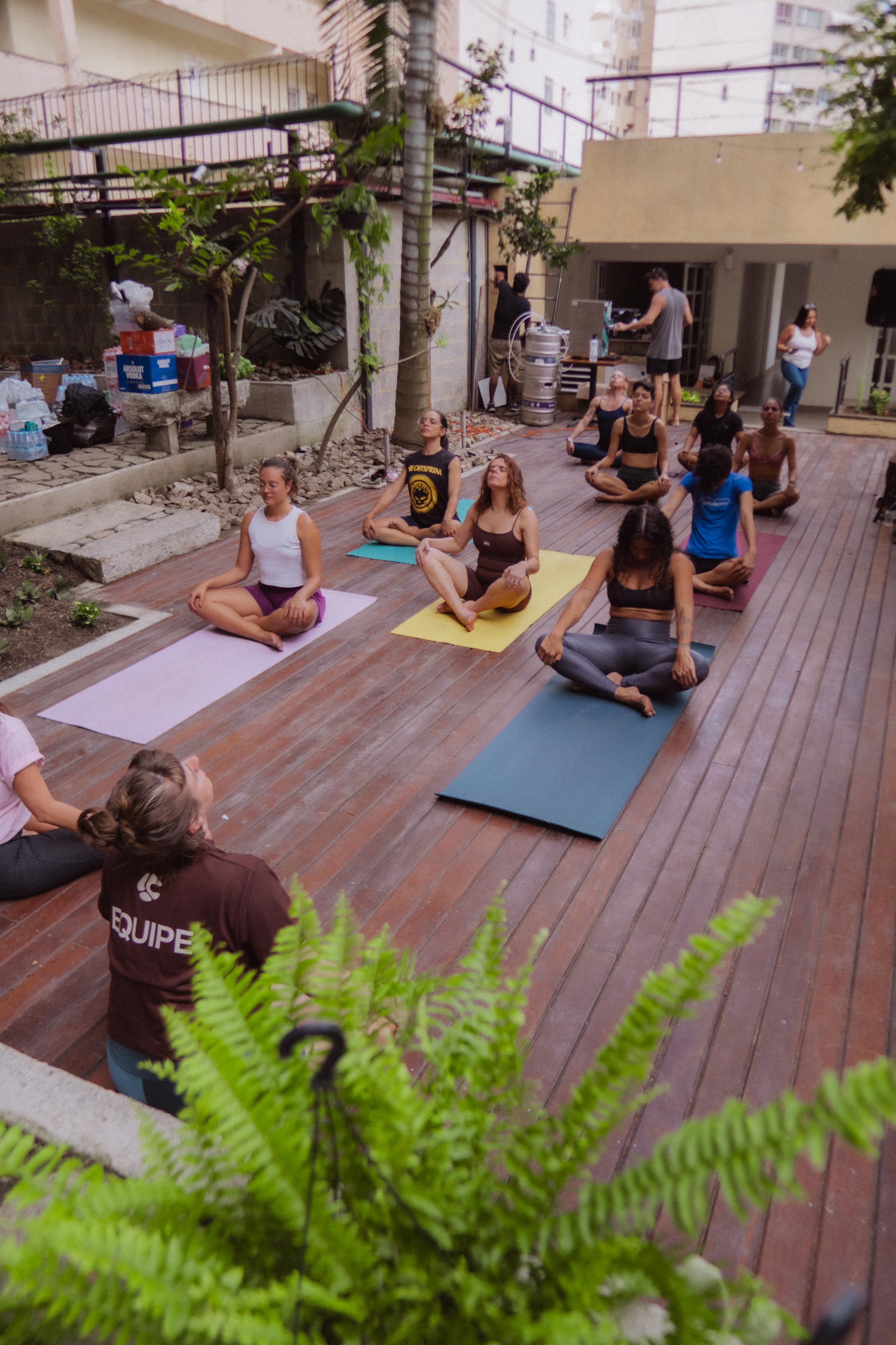 Roda de yoga no jardim: pessoas sentadas de pernas cruzadas em tapetes coloridos sobre o deck de madeira, com samambaias em primeiro plano e prédios ao fundo.